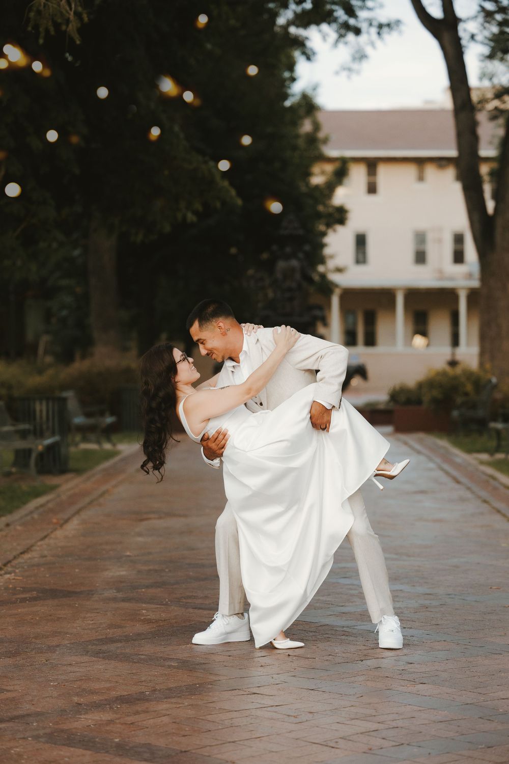 Bride and groom sharing a romantic dip kiss on a brick path at dusk with string lights and white venue behind.