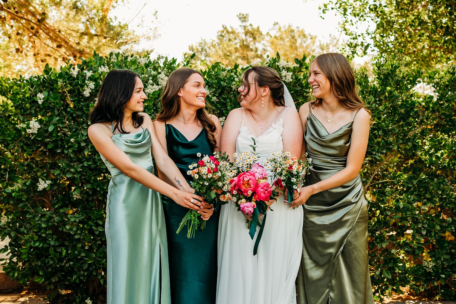 Bride in white gown with bridesmaids in green satin dresses laughing outdoors with pink floral bouquet.