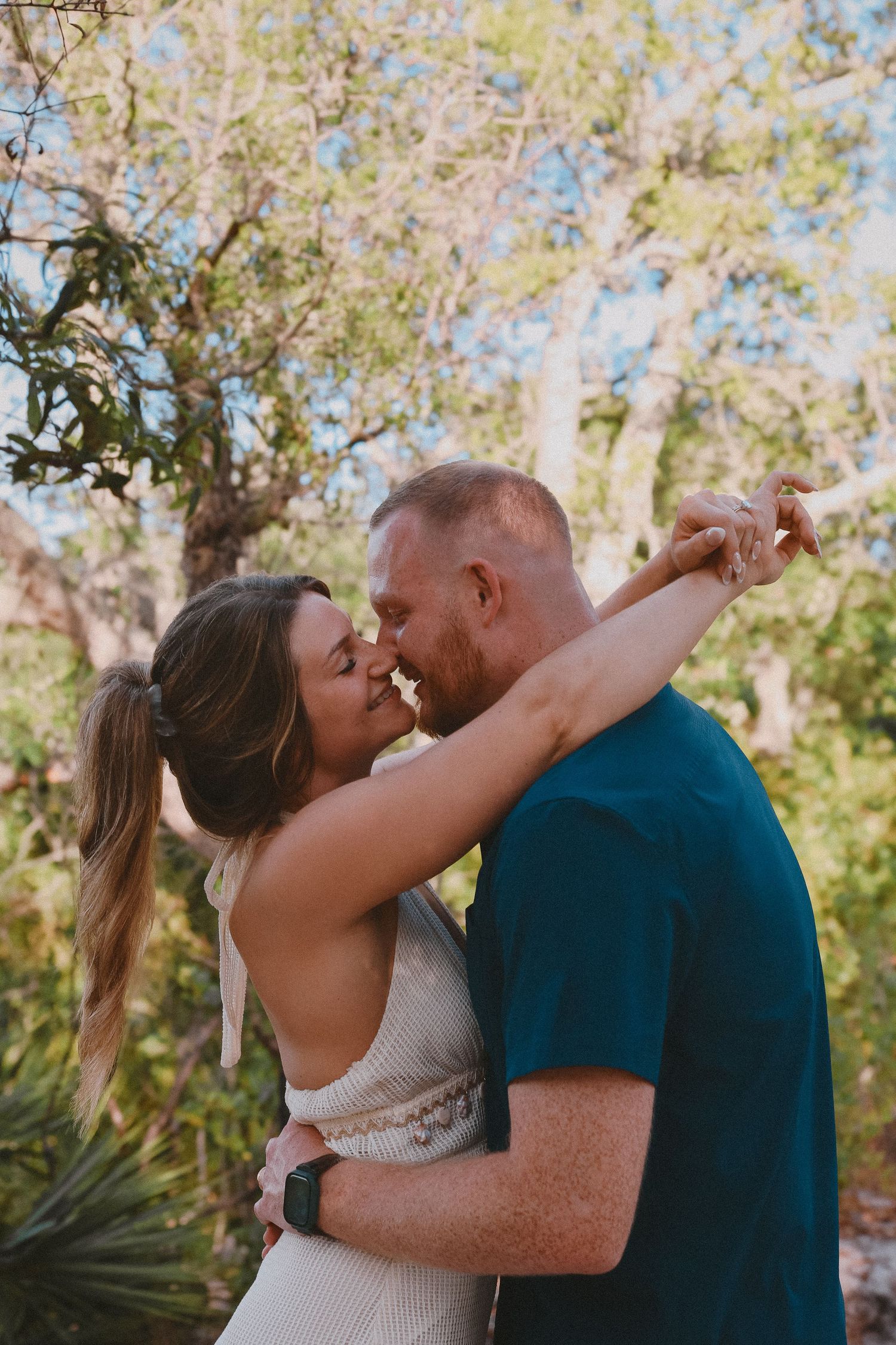 Couple sharing a kiss outdoors surrounded by lush green trees during engagement photo session.