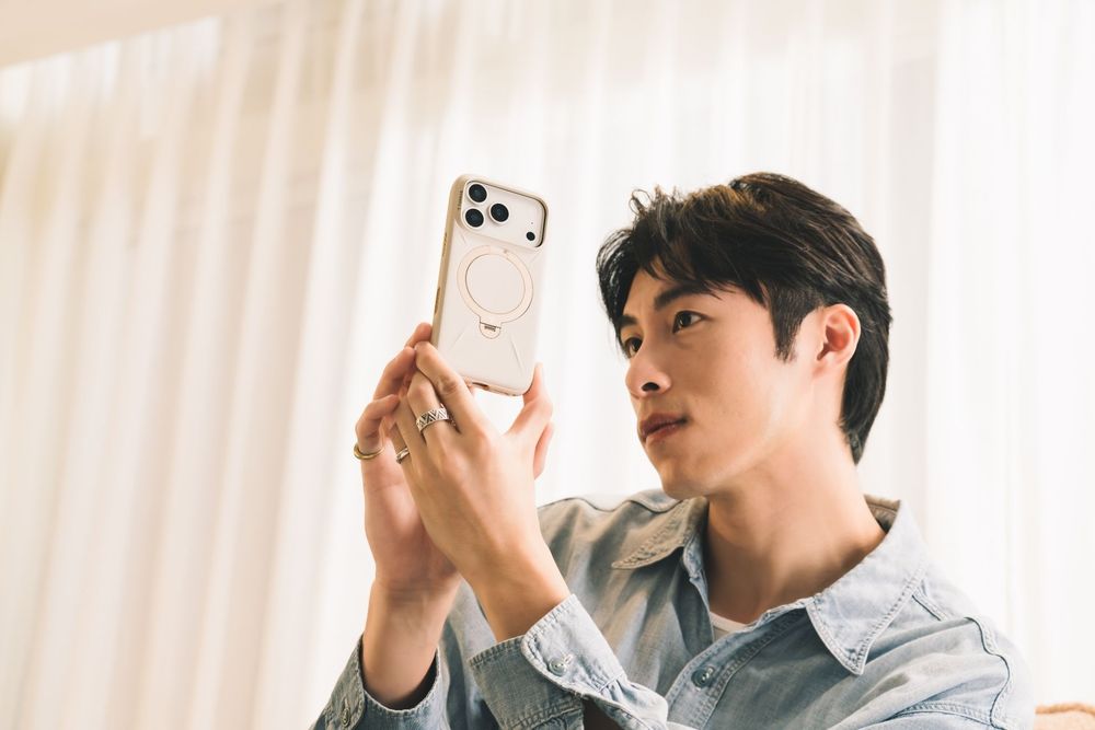 Young Asian man holding iPhone with clear case, taking a photo or video near a bright window.