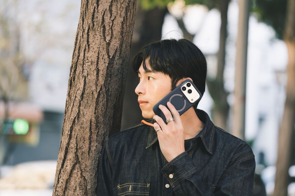 Young Asian man leaning against a tree trunk, talking on a smartphone outdoors in a denim jacket.