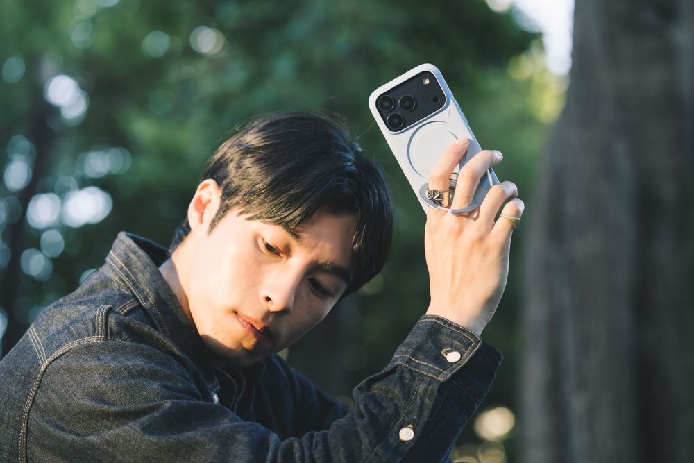 Young Asian man in dark jacket holds white smartphone up near his face outdoors among green trees.