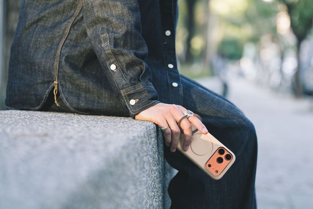 Person sitting on stone steps holding a smartphone with an orange MagSafe case, wearing dark denim outfit.