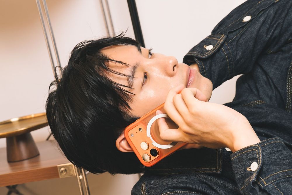 Young Asian man resting his chin on his hand while holding an orange phone case, sitting near a wooden desk.