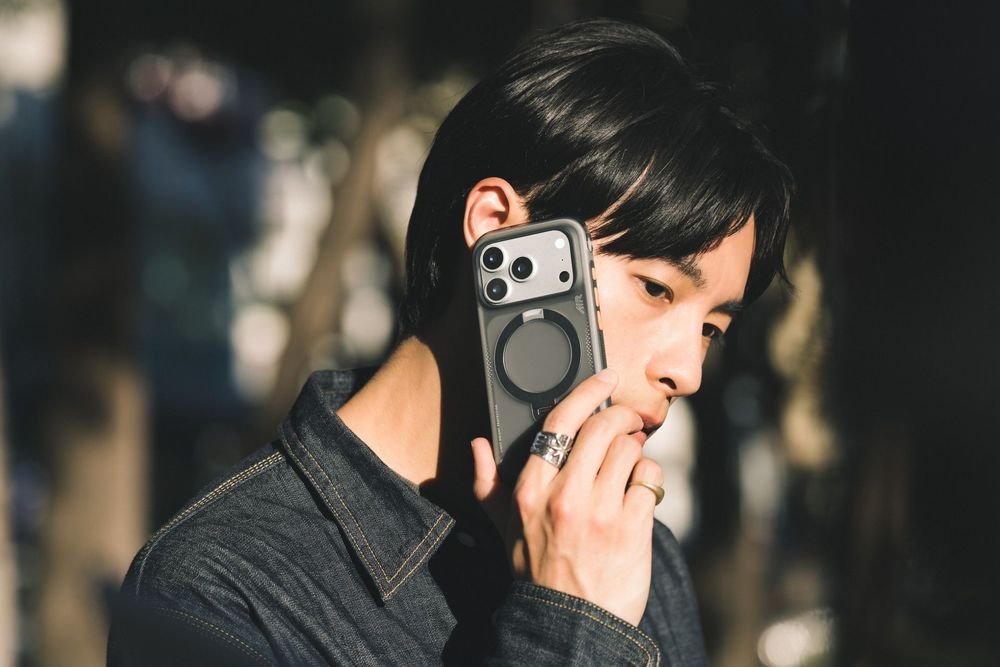 Young Asian man talking on a smartphone with a MagSafe case, wearing a denim jacket outdoors.