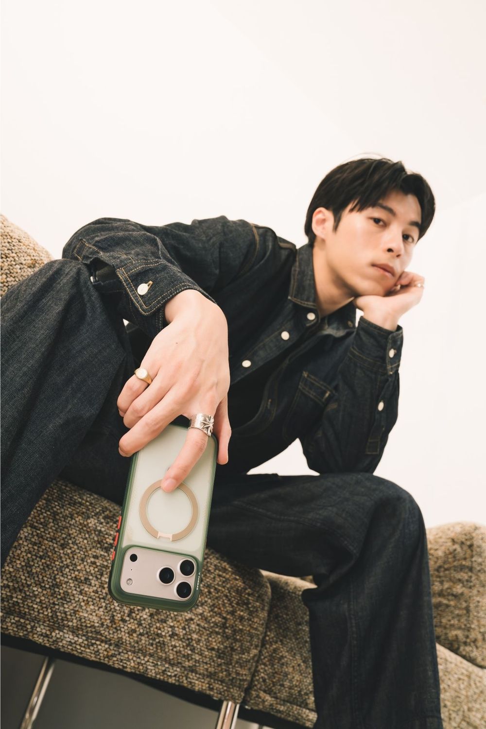 Young man in dark denim outfit sitting casually, holding a green phone case, wearing gold rings, with a relaxed pose.