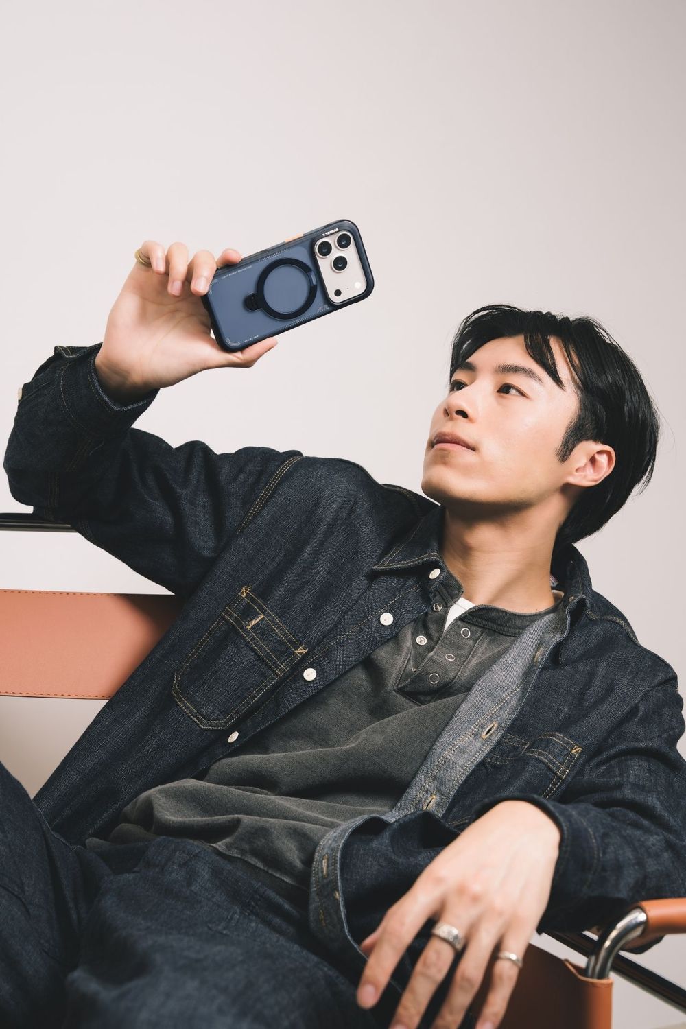 Young Asian man in denim outfit taking a selfie with a phone in a MagSafe case while seated.