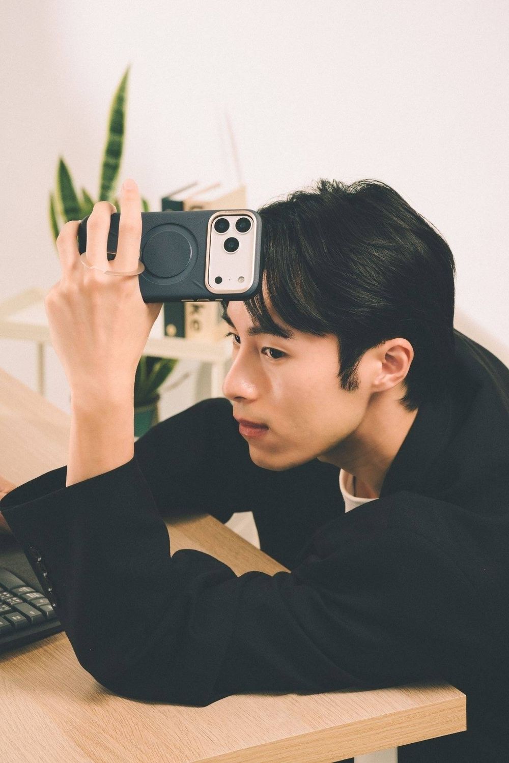 Young Asian man in black jacket examining a small camera closely while sitting at a wooden desk.