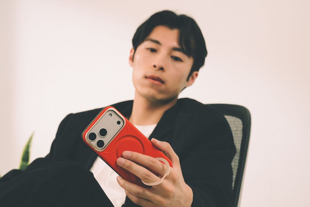 Young Asian man in black blazer holding a red iPhone case while sitting in an office chair.