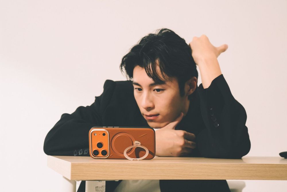 Young Asian man in black jacket resting on wooden table, looking at orange iPhone with ring holder.