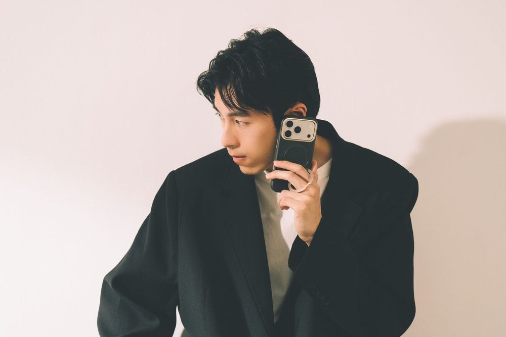 Young Asian man in black blazer holding smartphone against light background, stylish modern fashion portrait.