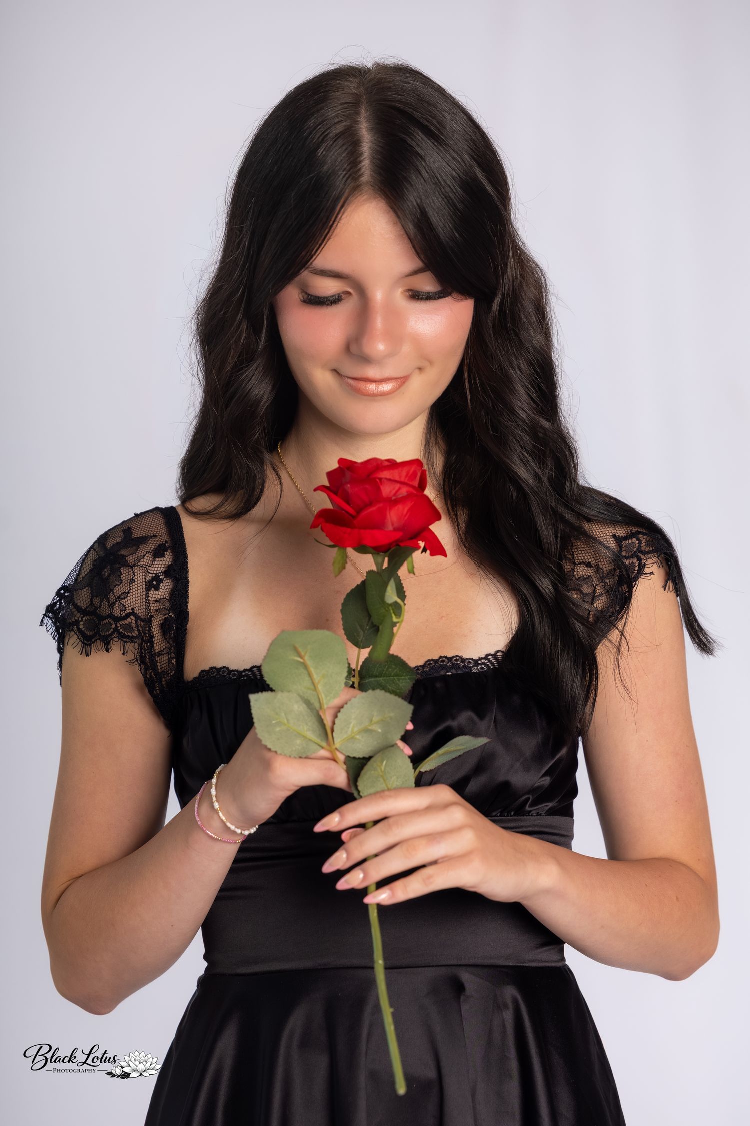 Young woman in black lace dress holding a red rose, looking down with a gentle smile, studio portrait.