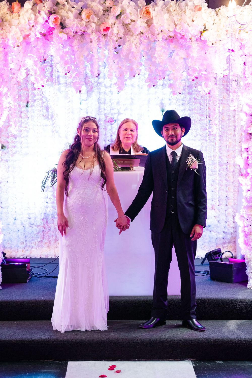 Bride and groom holding hands at wedding ceremony with floral backdrop, groom wearing black cowboy hat and suit.