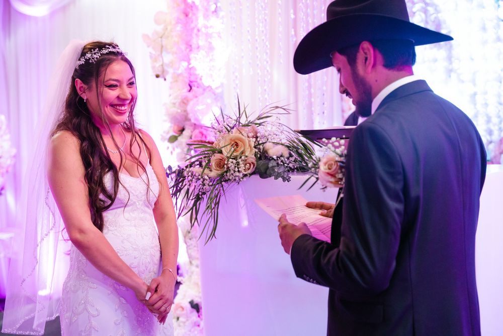 Bride smiling joyfully at groom reading vows during elegant wedding ceremony with floral backdrop and bouquet.