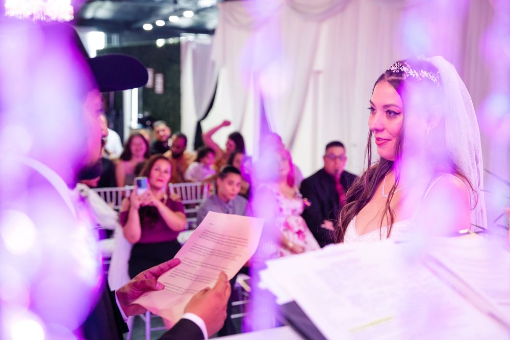 Smiling bride in tiara reads vows at elegant wedding reception with purple lighting and guests applauding in background.