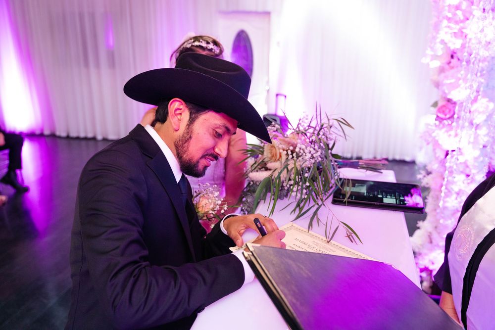 Jewish groom signing ketubah marriage contract at elegant wedding ceremony with purple lighting and floral decor.