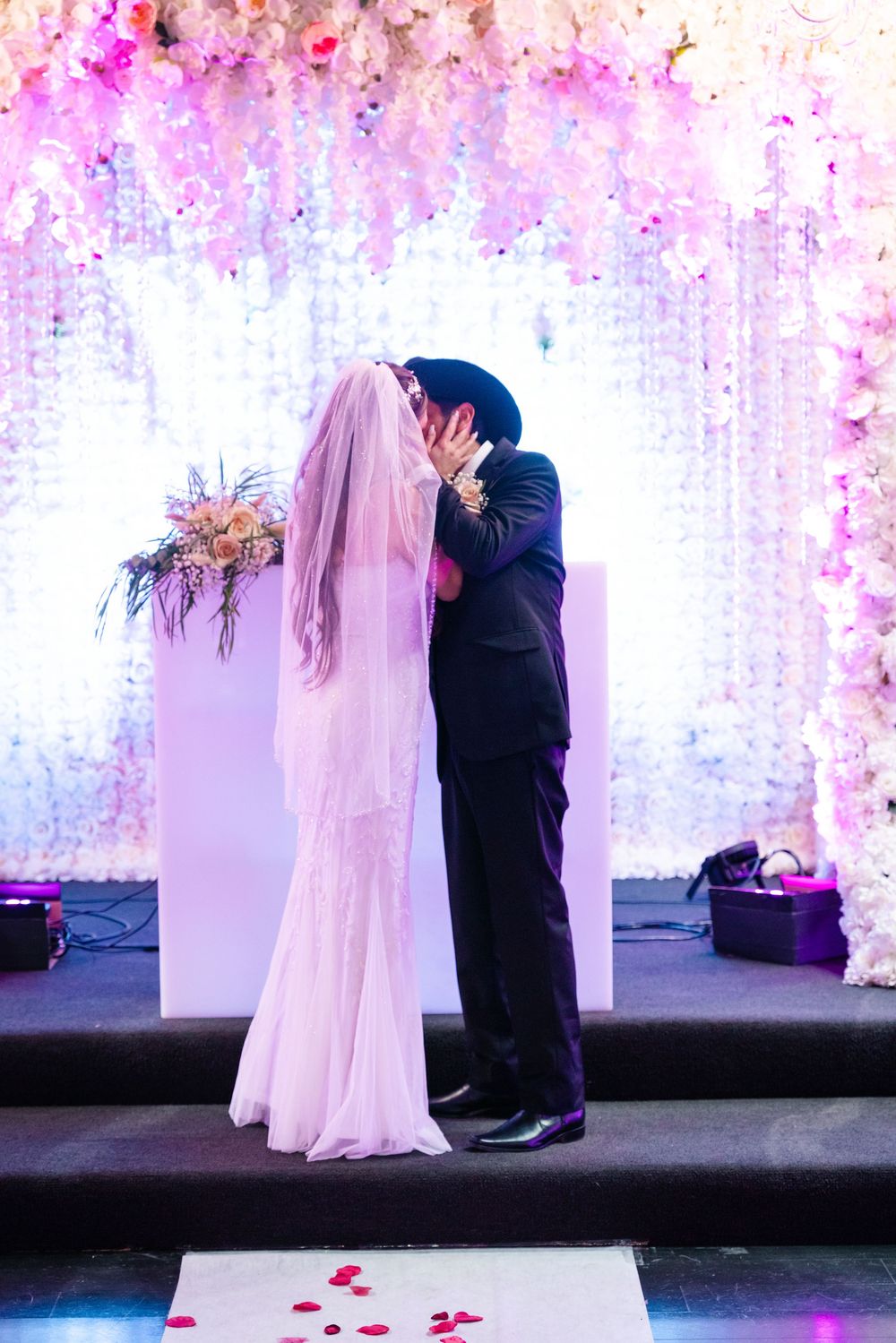Bride and groom share first kiss at altar adorned with cascading white and pink floral decorations at wedding ceremony.