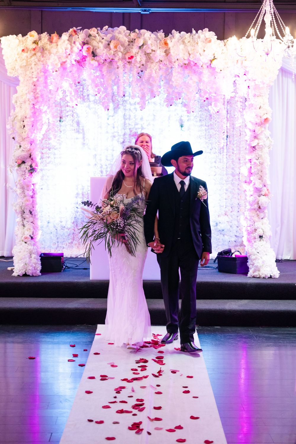 Bride and groom walk down rose petal aisle beneath stunning floral arch with cascading white flowers at wedding ceremony.