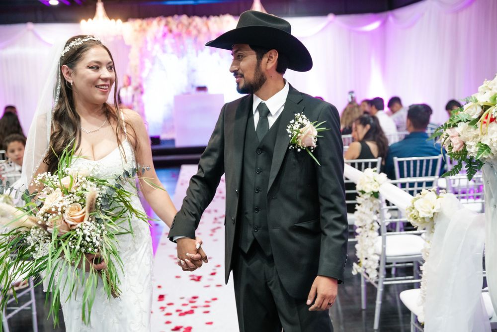 Bride and groom holding hands walking down the aisle at a wedding reception, groom wearing a black cowboy hat and suit.