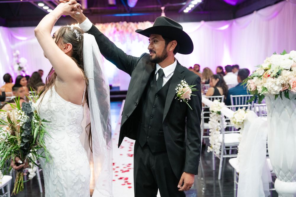 Bride and groom share first dance at wedding reception, groom wearing black suit and cowboy hat with boutonniere.
