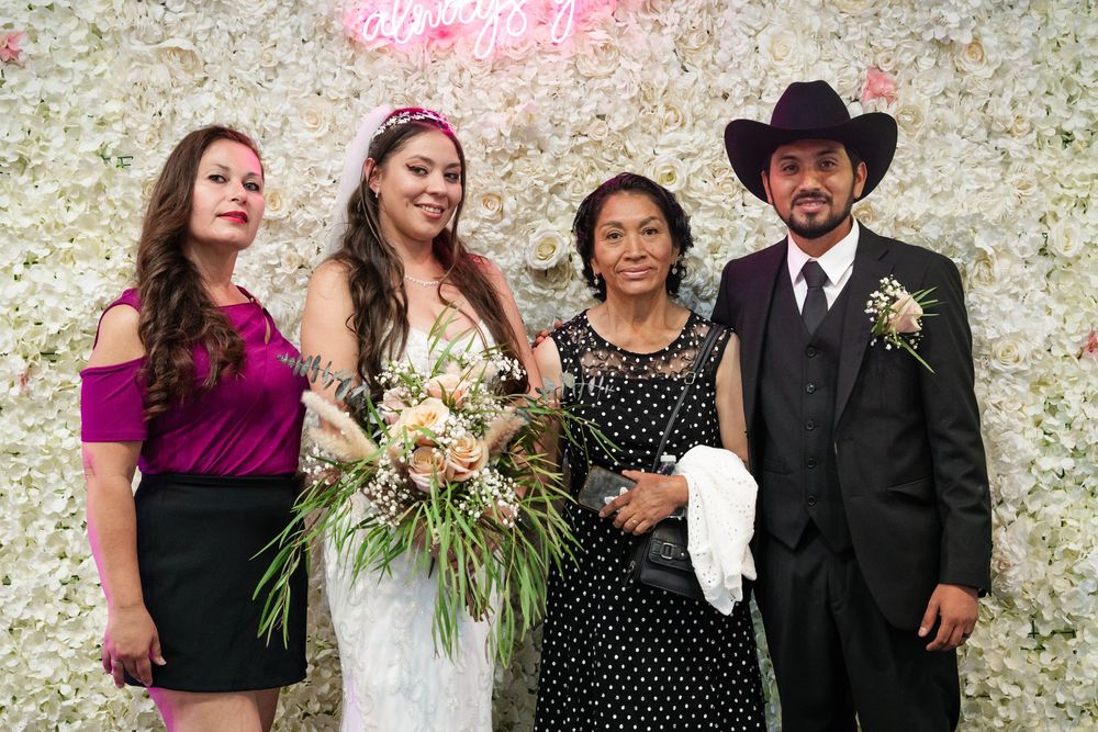 Bride in white gown holds bouquet with three guests at floral wall backdrop during wedding celebration.