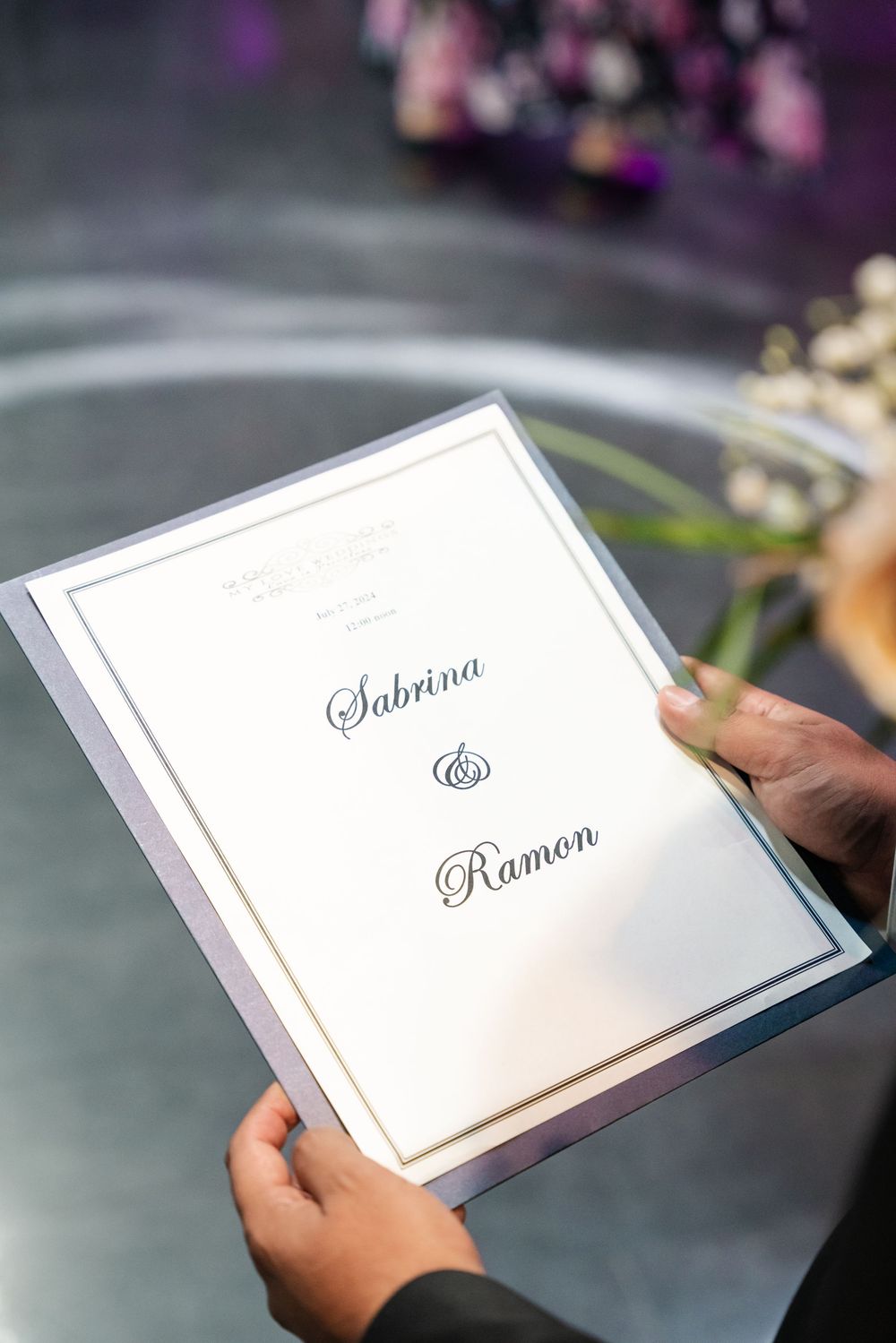 Hands holding a wedding program booklet with elegant script reading Sabrina and Ramon on white card with gold border.