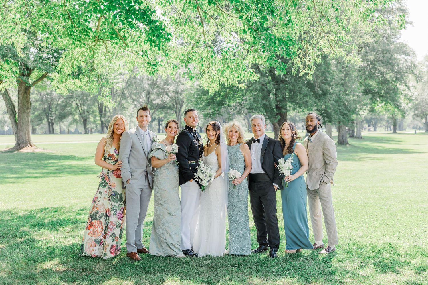 Wedding party poses outdoors in a lush green park, featuring bride and groom with family in elegant formal attire.