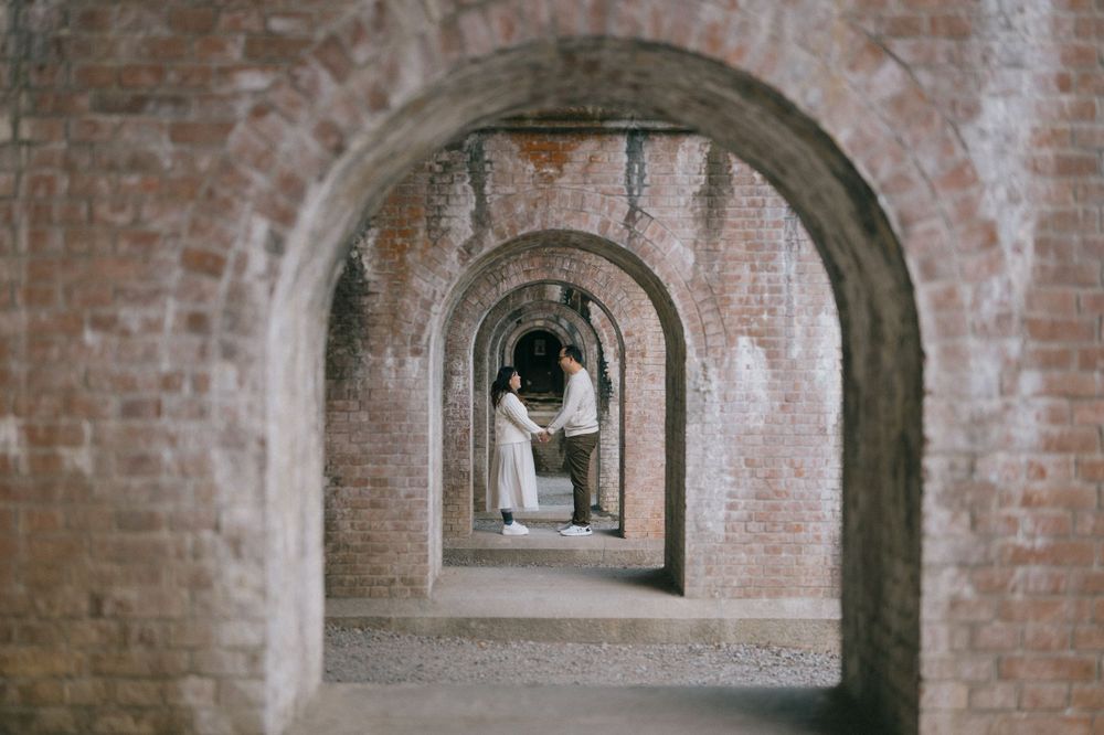 Couple holds hands framed by a series of rustic brick archways creating a romantic tunnel perspective.
