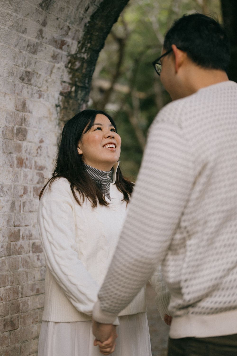 Smiling Asian woman looks up lovingly at her partner while holding hands near a rustic brick wall outdoors.