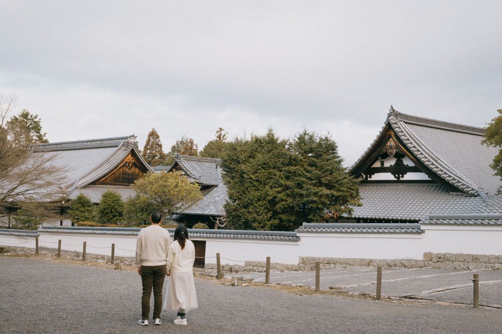 Couple standing together viewing traditional Japanese temple buildings with tiled roofs on overcast day.