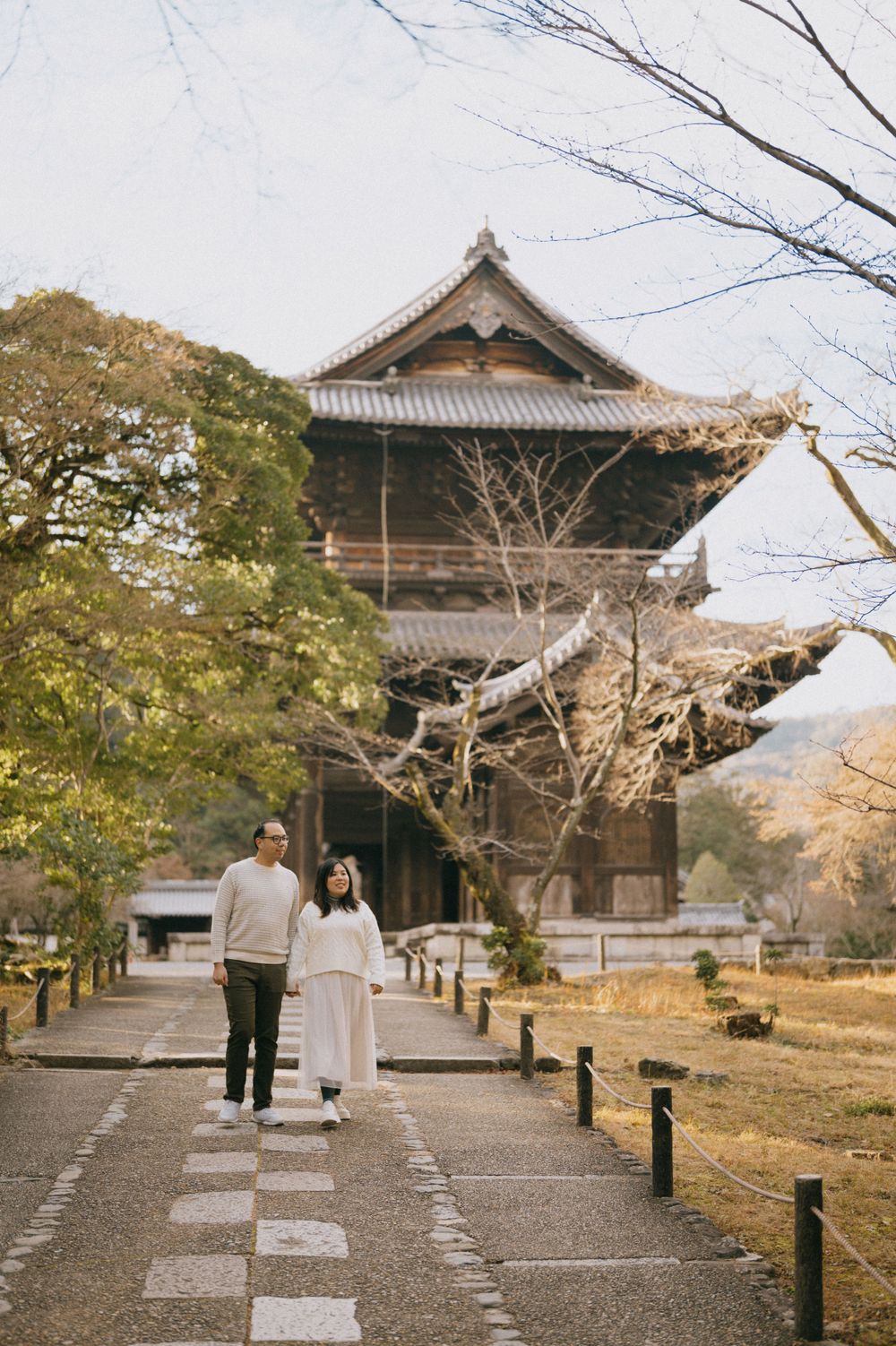 Couple standing on checkered path before large traditional Japanese wooden pagoda with bare winter trees.