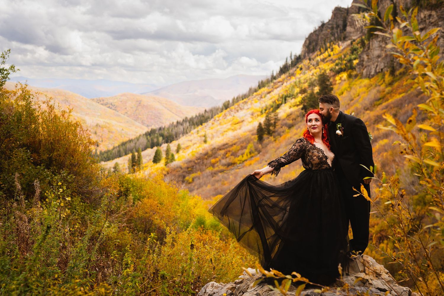 Couple posing on a rocky mountain overlook surrounded by vibrant fall foliage.