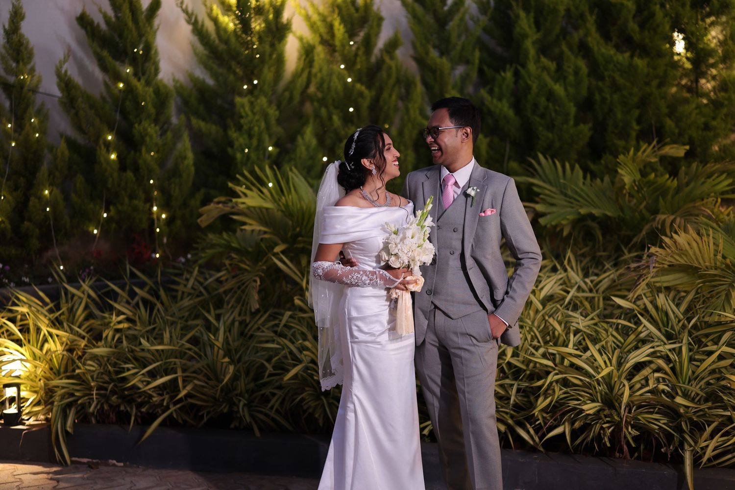 Elegant bride and groom in wedding attire standing together outdoors surrounded by lush green plants at night.