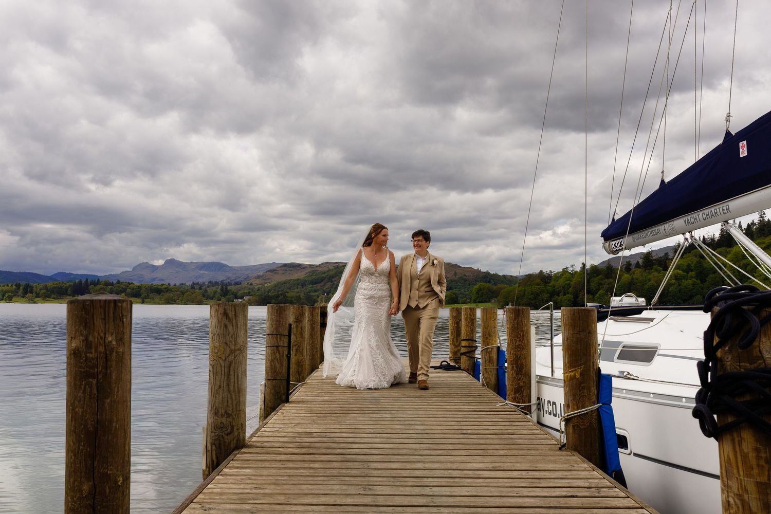 Two brides walk hand in hand on a wooden dock by a lake, with sailboats and dramatic cloudy skies behind them.