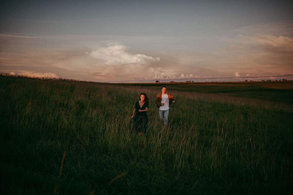 Two people walking hand in hand through a lush green field at sunset under a dramatic cloudy sky.