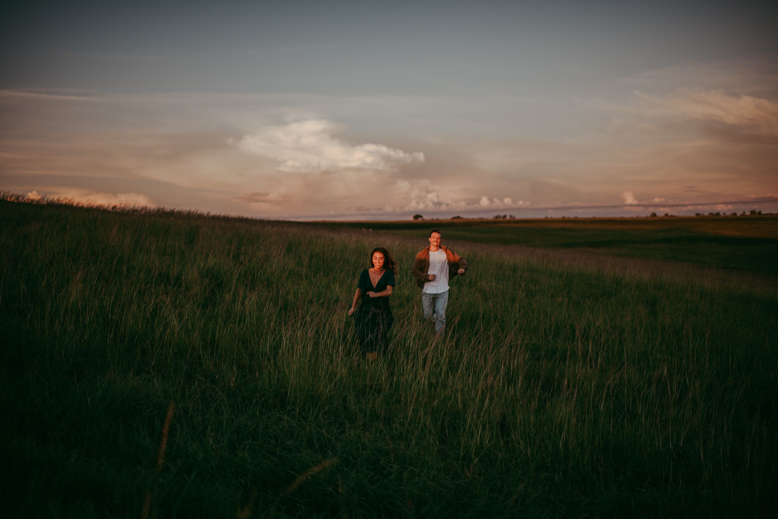Two people walking hand in hand through a lush green field at sunset under a dramatic cloudy sky.