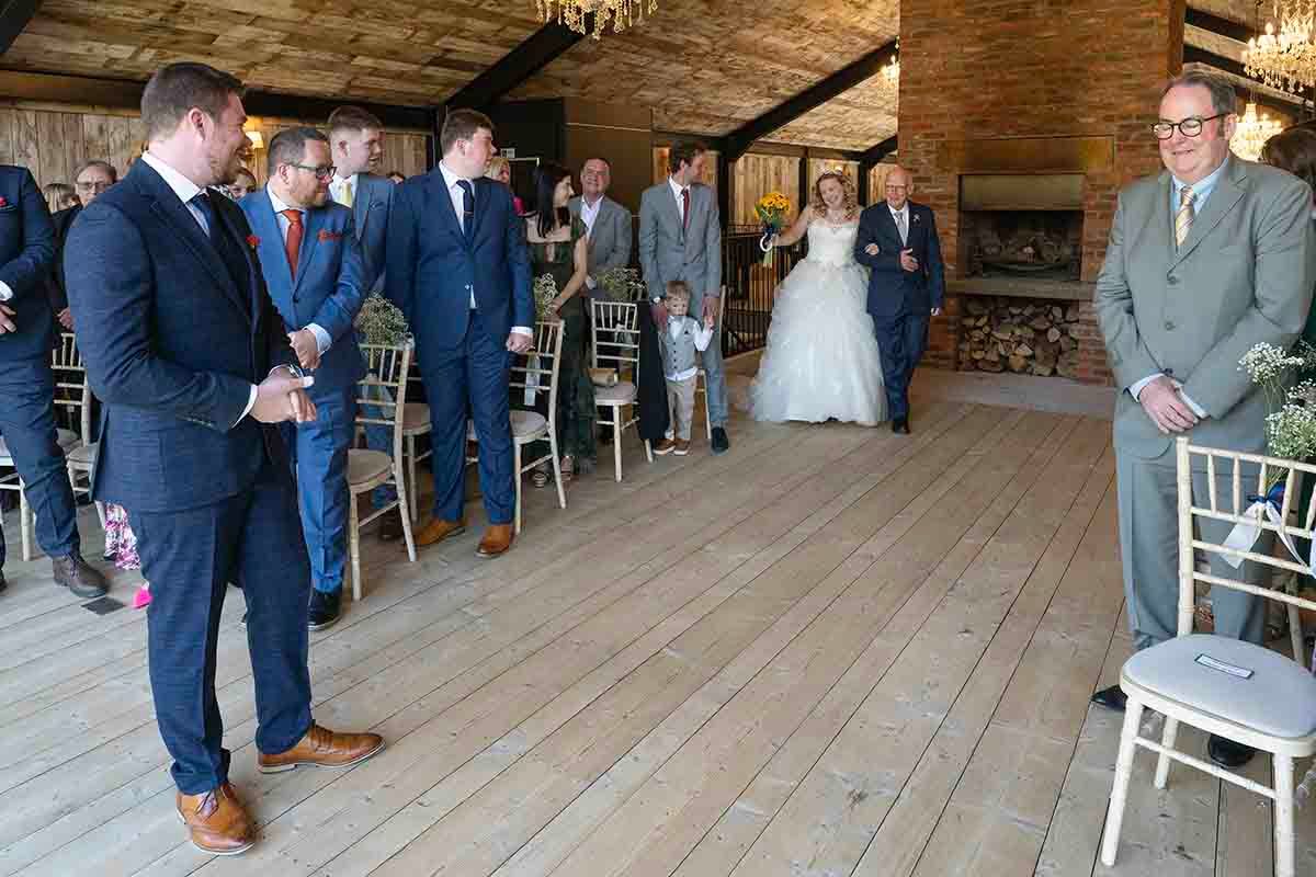 Bride walking down the aisle with escort at a rustic barn wedding ceremony with groomsmen waiting.
