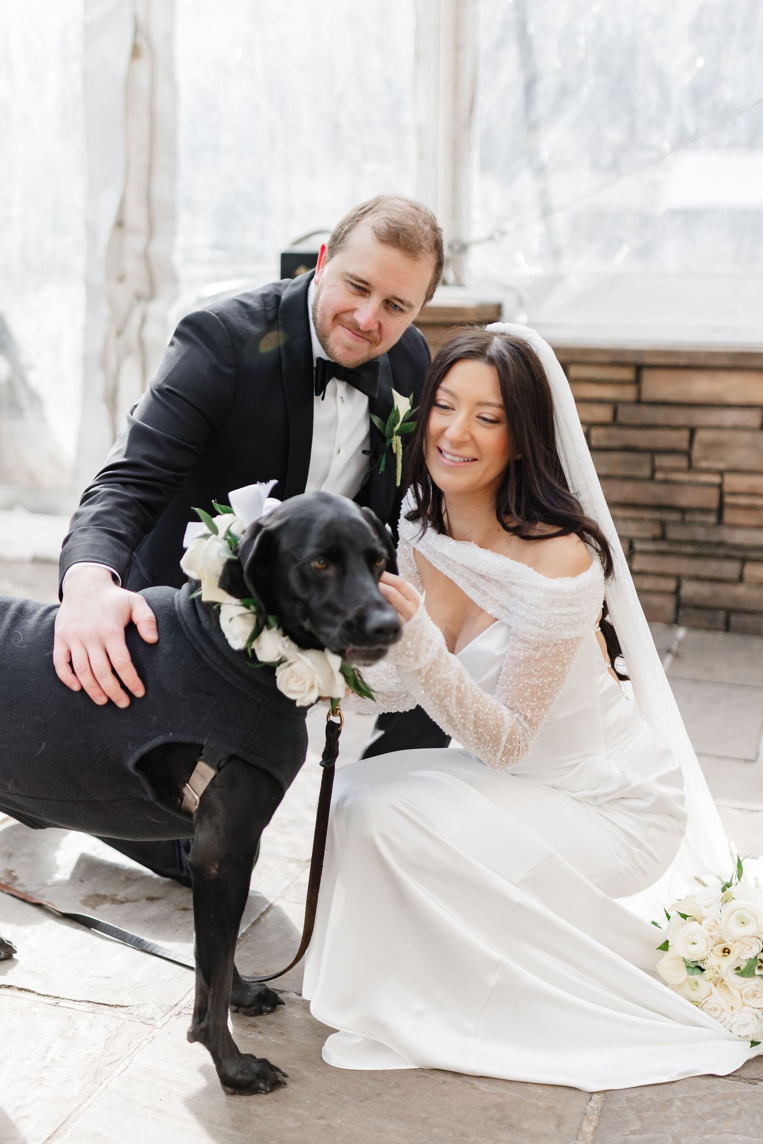 Bride and groom in wedding attire smile while petting their black Labrador dog wearing a white floral collar.