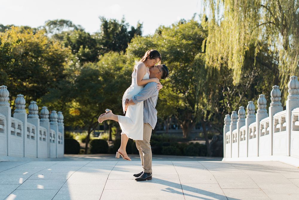 Couple sharing a romantic embrace on a sunlit bridge surrounded by lush green trees in a park. photoshoot