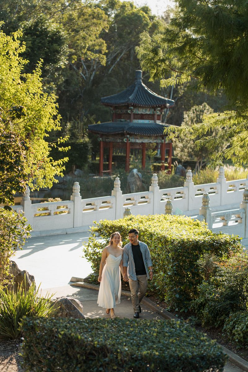 Couple holding hands walking through a Chinese garden with a traditional red pagoda and white stone bridge surrounded by lush greenery.