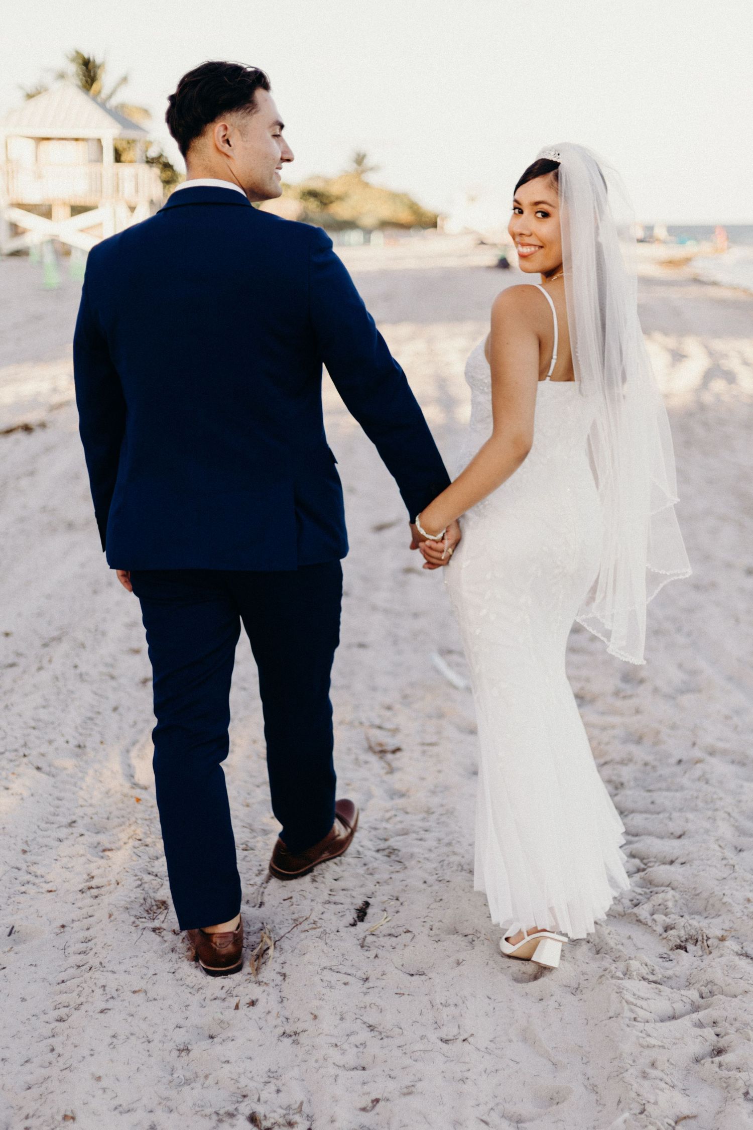 A couple in wedding attire holding hands while walking on a sandy beach at sunset.