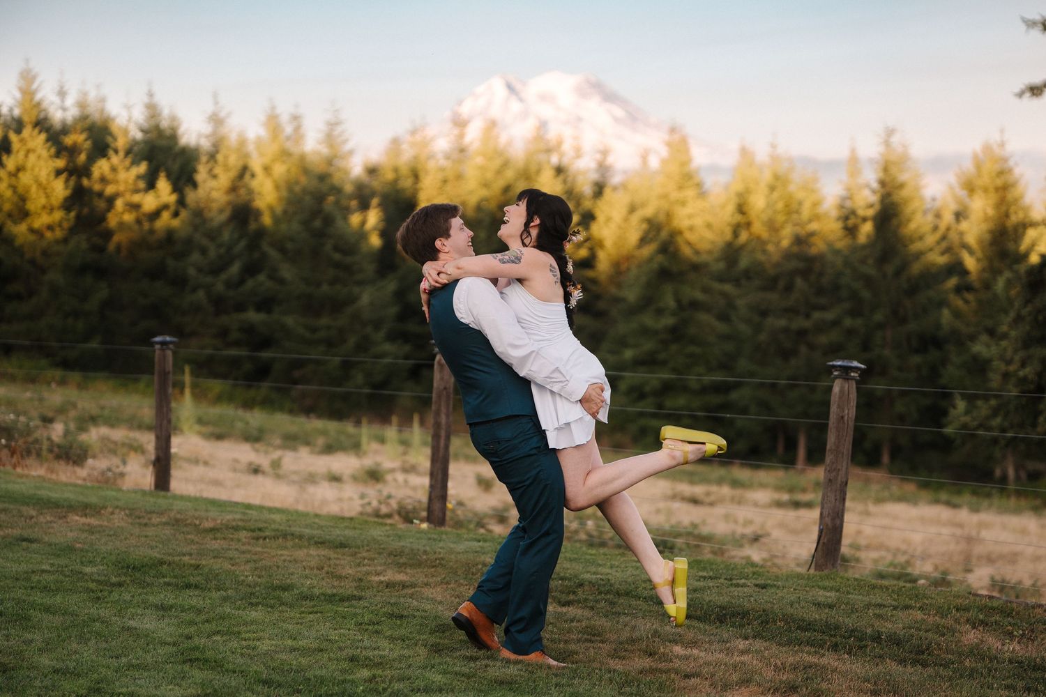 Mackie lifting Briana in white dress with Mount Rainer and pine trees in golden hour background at their Winter View Lodge Wedding.