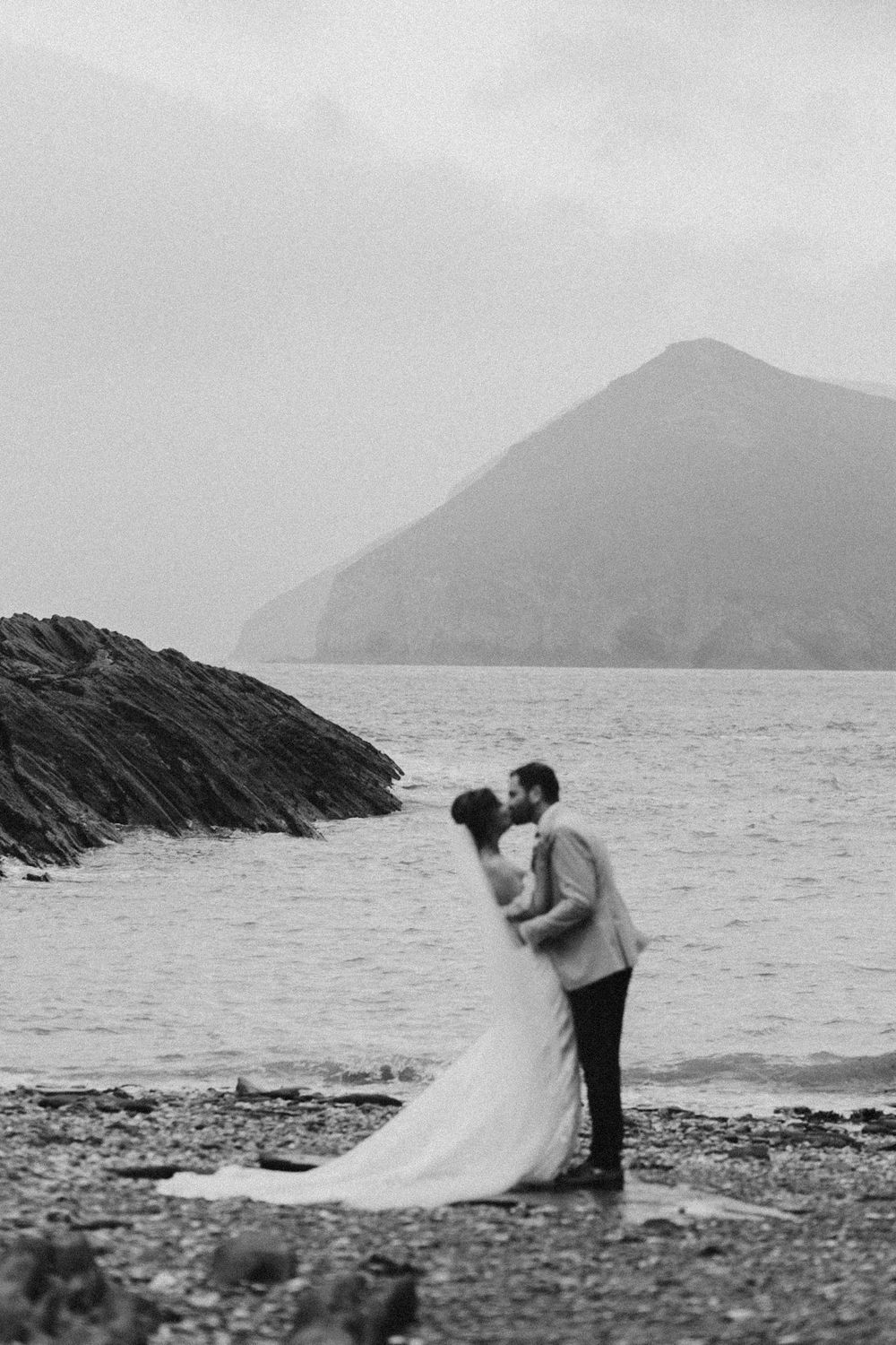 Wedding couple embraces on rocky shoreline with misty mountain and ocean backdrop.