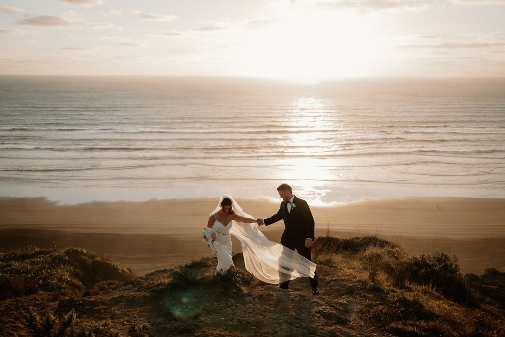 Two silhouettes stand on a coastal cliff at sunset while a veil flows dramatically in the ocean breeze.