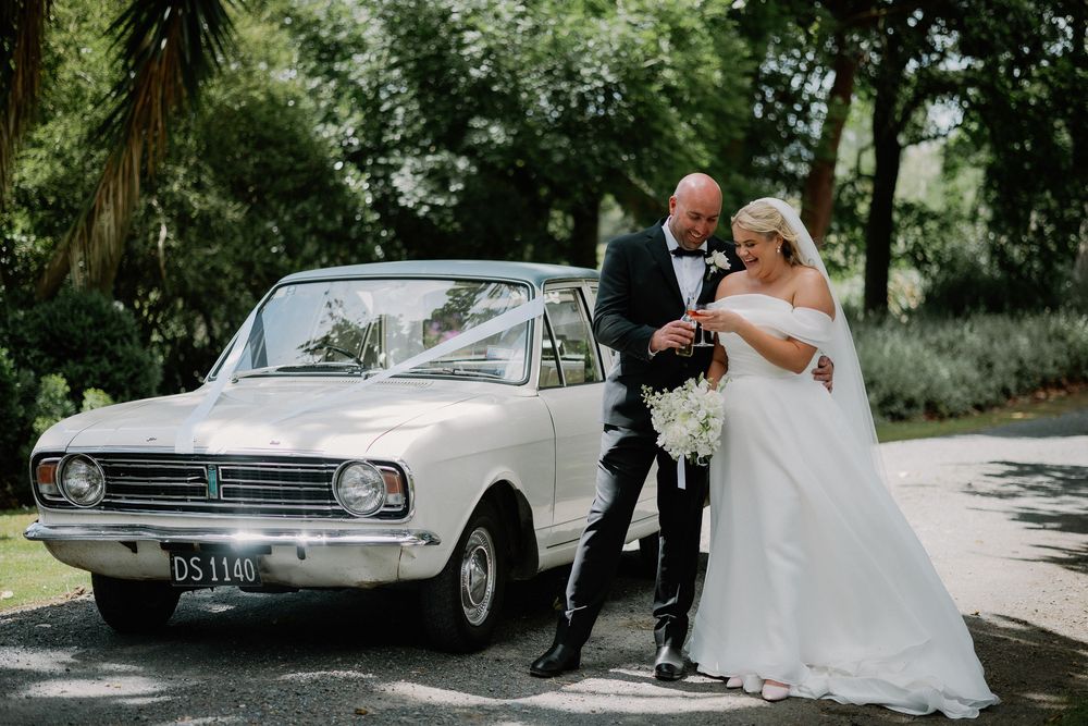 Bride and groom posing beside a classic white vintage car decorated with ribbon on their wedding day.