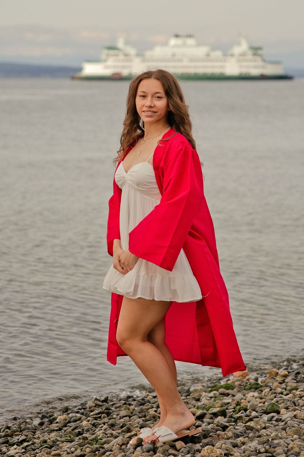 Young woman in red robe and white dress poses on rocky beach with ferry boat on calm water behind her.