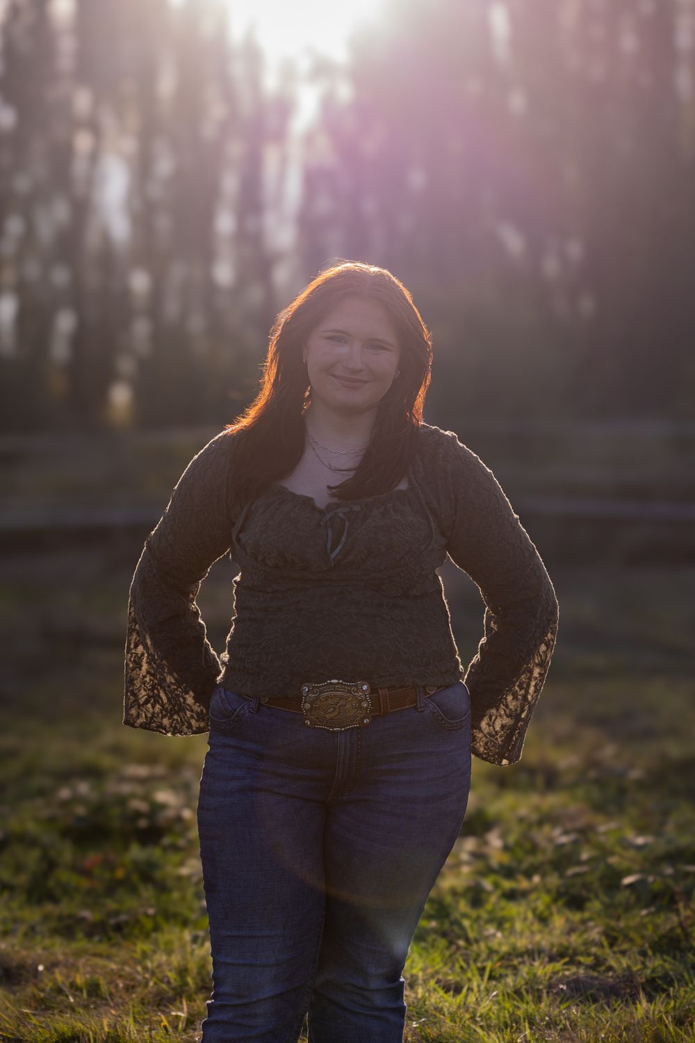 Woman in dark top and jeans with belt buckle smiling outdoors in golden sunlight with trees in background.