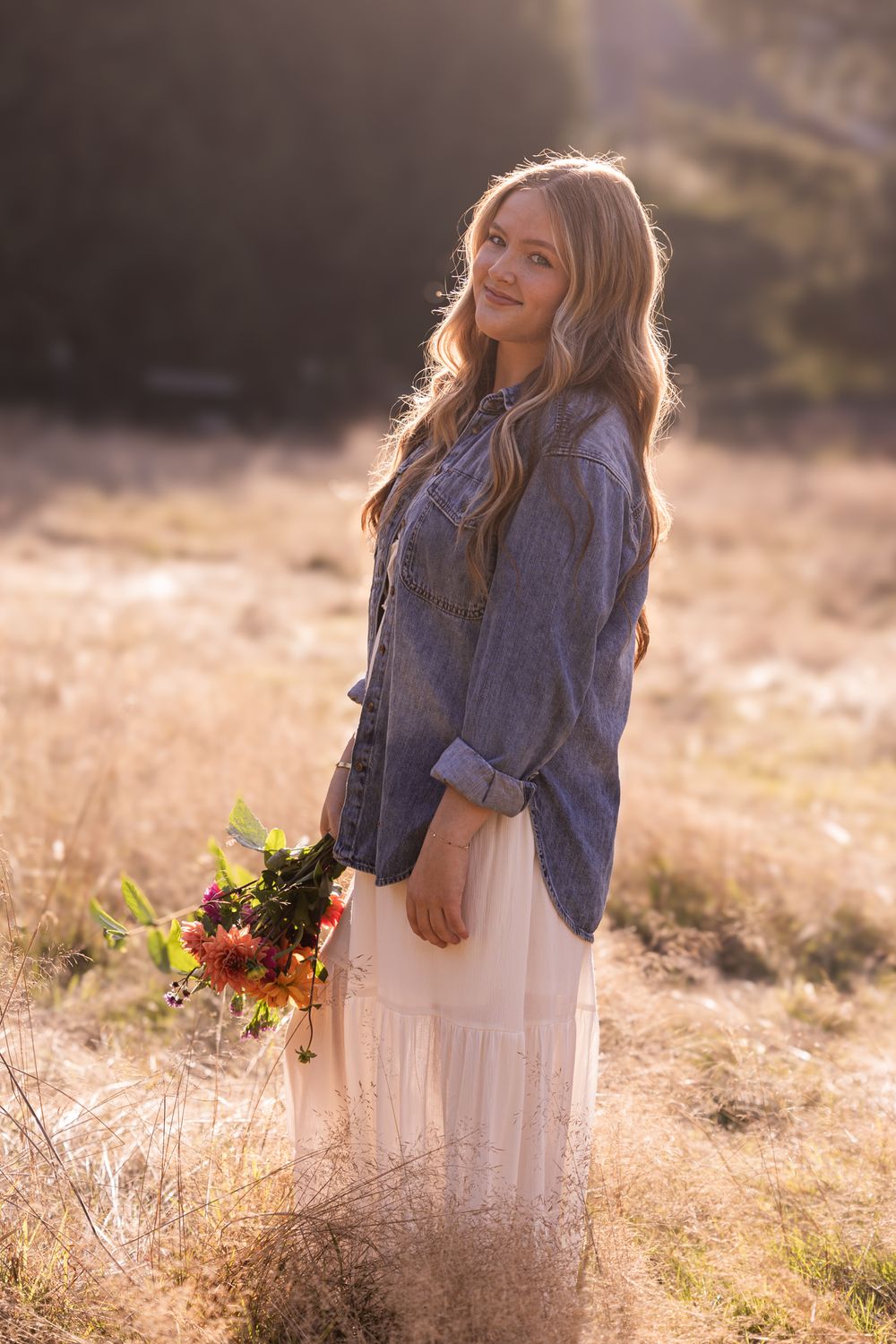 Young woman in denim shirt and white skirt holding wildflower bouquet in golden sunlit field.