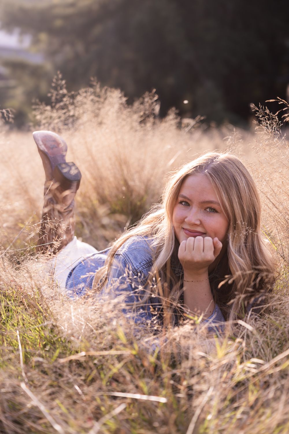 Young blonde woman lying in golden meadow grass, resting chin on hand, wearing denim jacket and boots in warm sunlight.