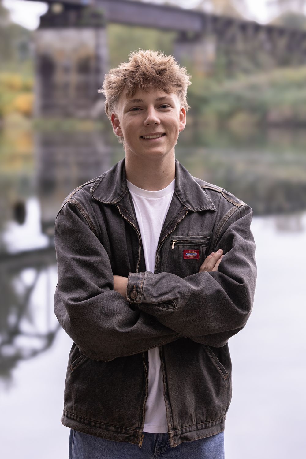 Smiling teenage boy with blonde hair, arms crossed, wearing a dark denim jacket near a river bridge.